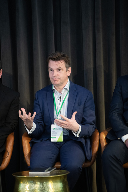 Three men in business attire sit in a row, the central figure speaking on stage into a microphone that is clipped to his b...